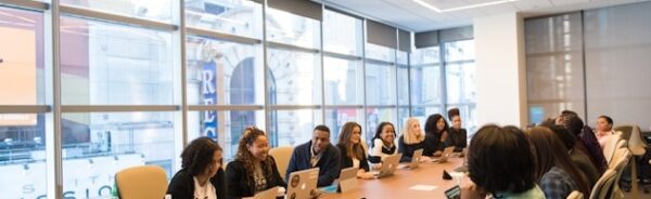 Diverse group of professionals seated around a long conference table with laptops in a bright modern office meeting room.