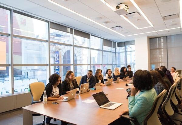 Diverse group of professionals seated around a long conference table with laptops in a bright modern office meeting room.