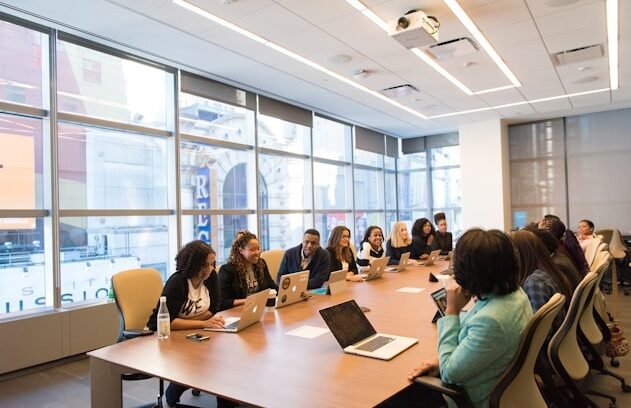 Diverse group of professionals seated around a long conference table with laptops in a bright modern office meeting room.